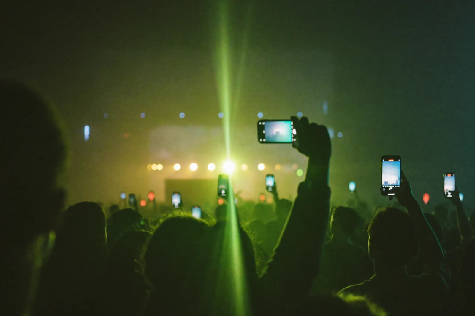 Un público de concierto de noche con teléfonos en alto hacia el escenario, atravesado por un destello de luz verde lima.