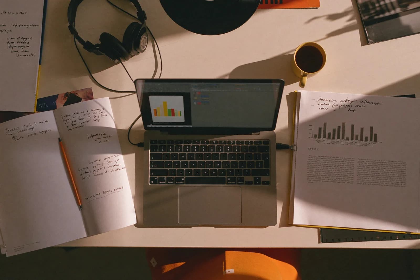 Un escritorio con luz de mañana: laptop, café, páginas impresas con gráficas y audífonos.