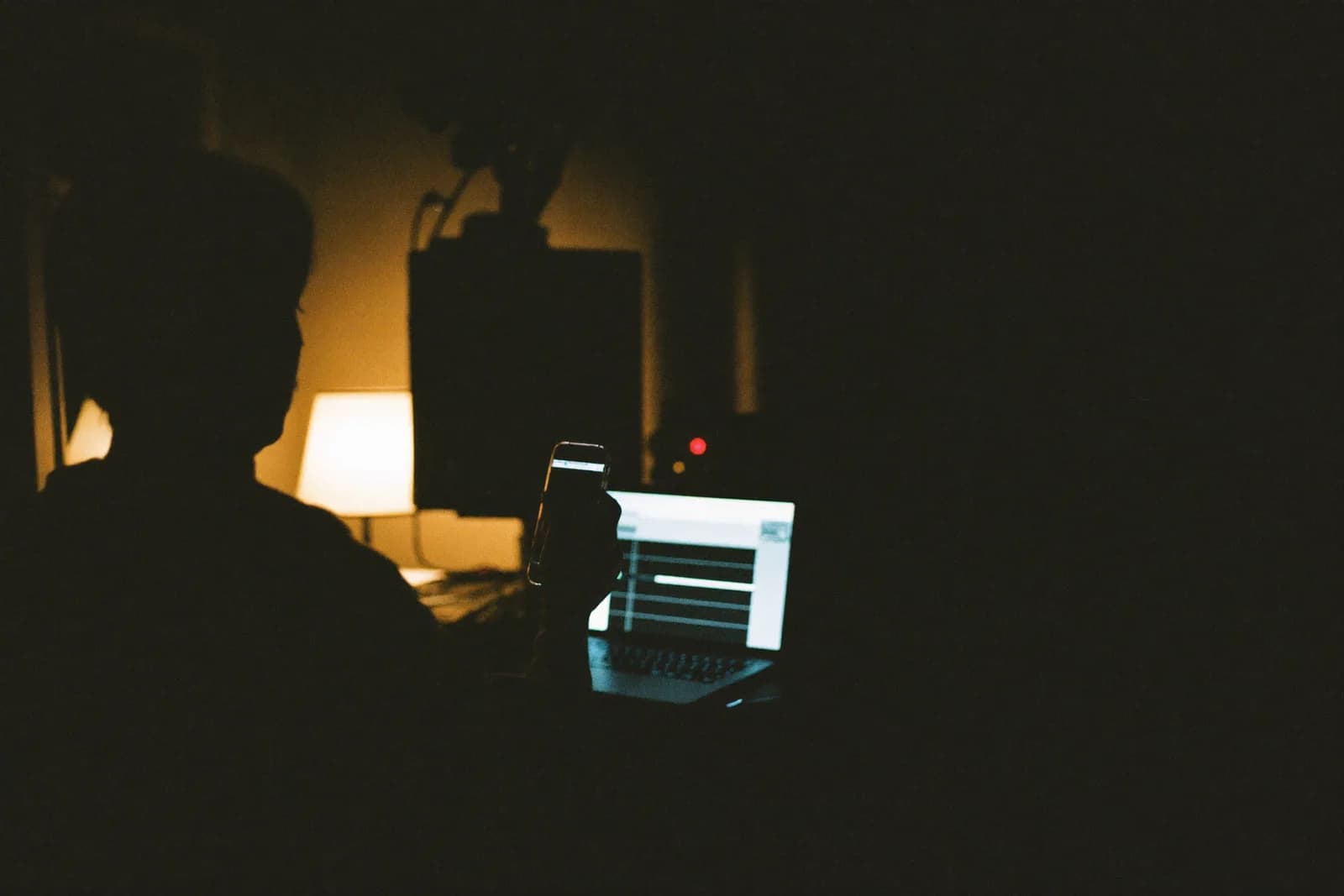 Un músico solo en un estudio casero de noche, teléfono en mano, con la laptop encendida al lado.